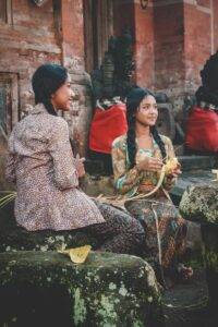 woman holding woven coconut leaf beside woman sitting while holding knife